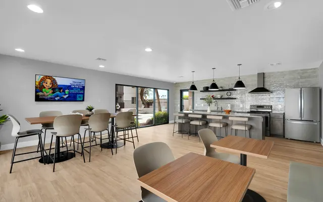 A modern kitchen and dining area featuring tables and chairs, a large window with natural light, a television on the wall, and a stylish kitchen counter with appliances.
