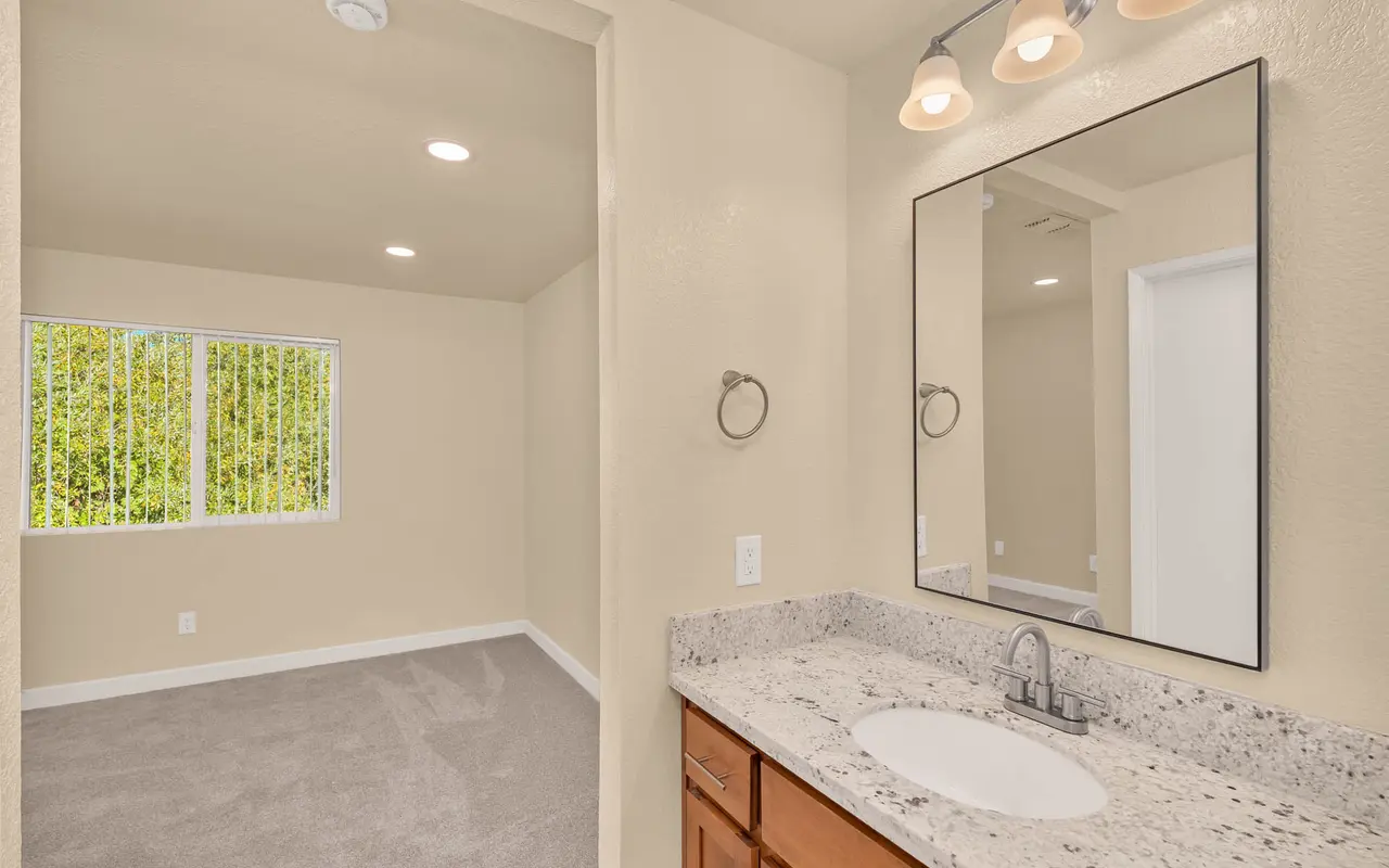 A view of a modern bathroom with a large mirror and granite countertop, leading into a bedroom with carpet and large window.