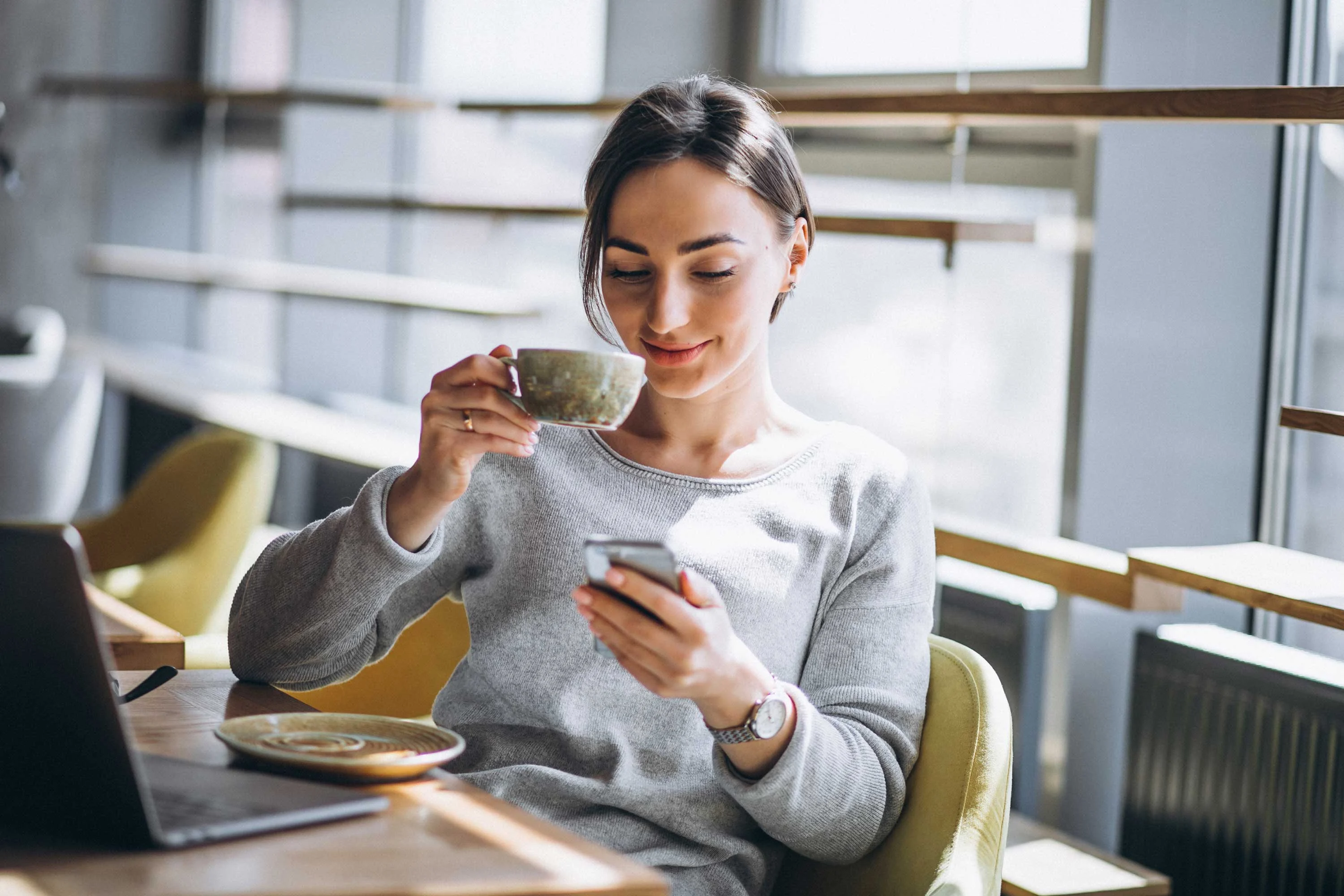 A woman sitting at a table in a cafe, holding a cup of coffee and looking at her smartphone, with a laptop and a plate nearby.
