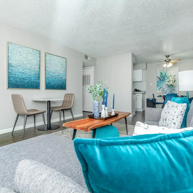 A modern living room featuring a grey couch adorned with colorful cushions, a wooden coffee table, and a small dining area with two chairs. The walls are decorated with blue-themed artwork, and there's a small kitchen visible in the background.