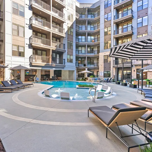 A modern apartment complex featuring a central swimming pool surrounded by lounge chairs and umbrellas, with a multi-story building in the background.