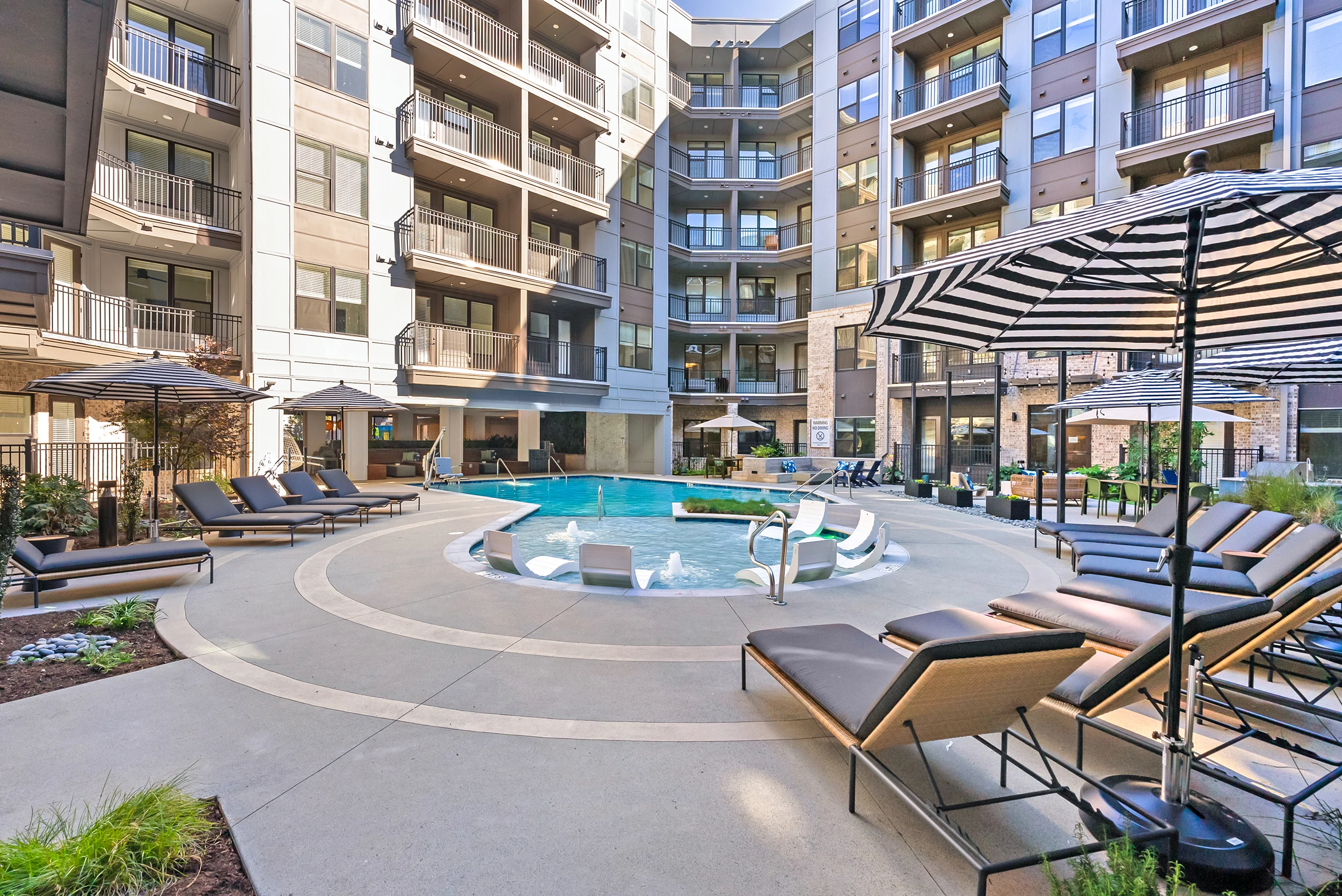 A modern apartment complex featuring a central swimming pool surrounded by lounge chairs and umbrellas, with a multi-story building in the background.