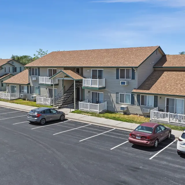 Apartment Complex View A two-story apartment complex with a tan-colored exterior and a brown roof. The building has multiple balconies and is surrounded by a parking lot with several cars parked. There are trees in the background and a clear blue sky overhead.