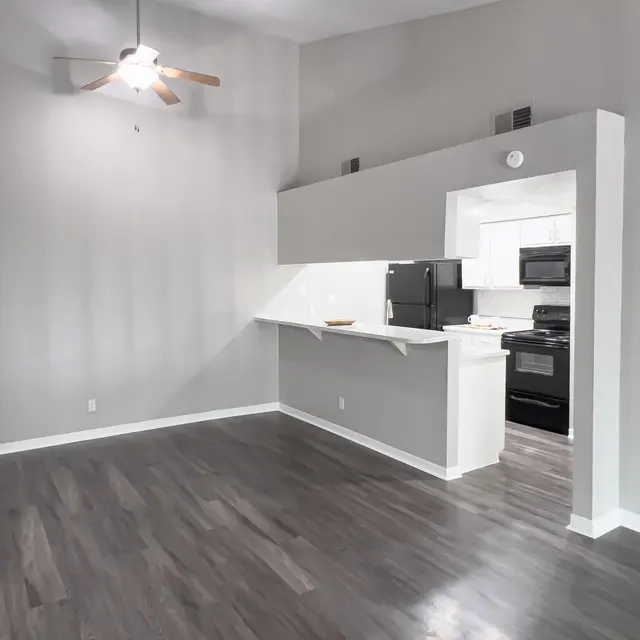 Spacious room with gray walls, wood-style flooring, ceiling fan, and a kitchenette featuring black appliances in the background.