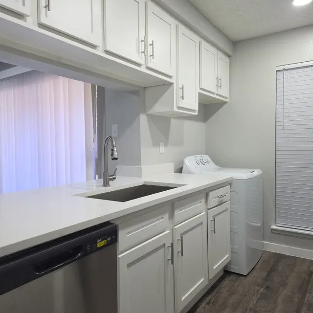 A modern kitchen featuring white cabinets, a stainless steel dishwasher, a sleek countertop, and a washing machine located beside the sink. There is natural light coming in from a window with white blinds.