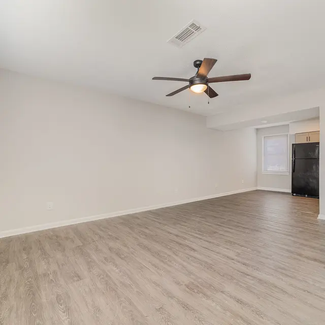 An empty living room with light-colored walls and a wooden floor. A ceiling fan is visible, and there is a portion of an adjoining kitchen with a black refrigerator in the background.
