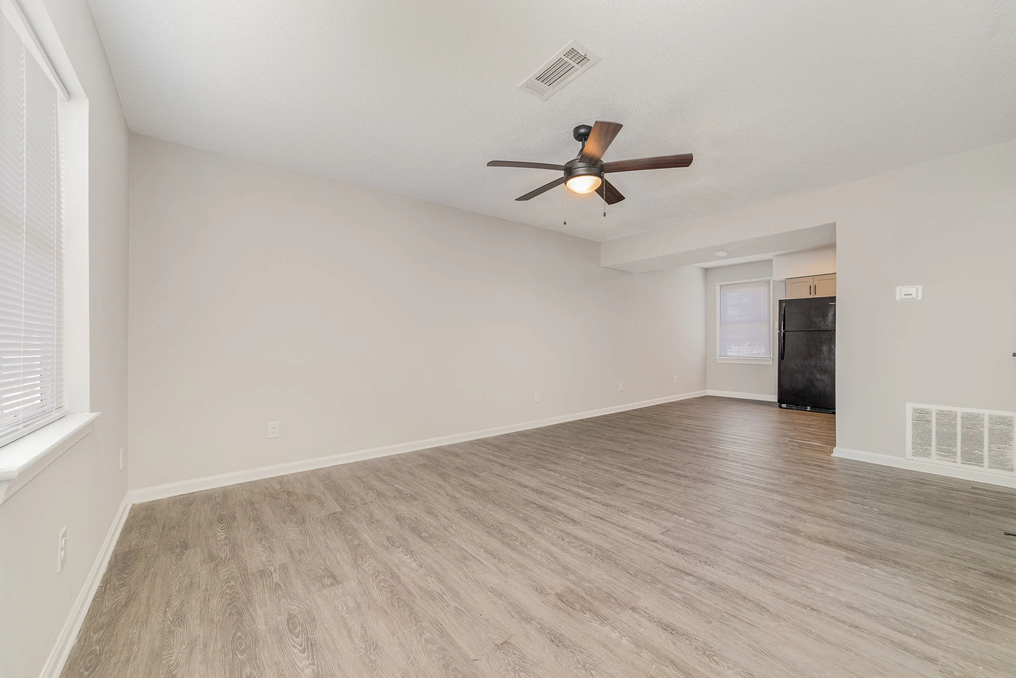An empty living room with light-colored walls and a wooden floor. A ceiling fan is visible, and there is a portion of an adjoining kitchen with a black refrigerator in the background.