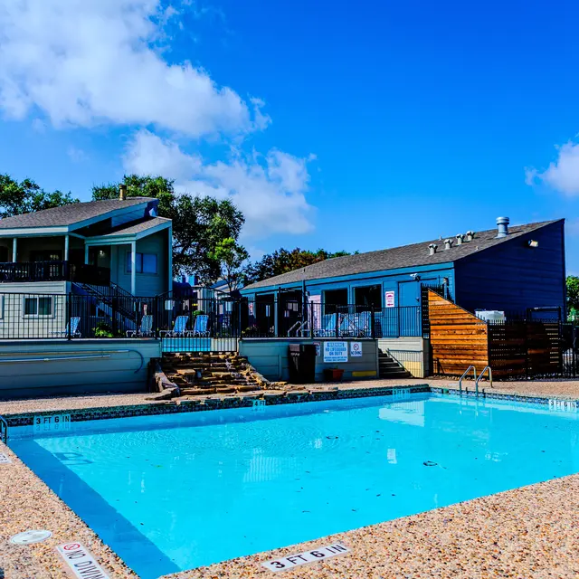 A sunny outdoor pool area with a clear blue sky, surrounded by two multi-story residential buildings and lounging chairs.