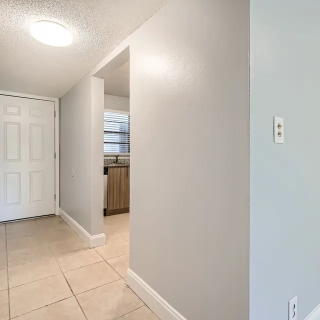 A hallway with light gray walls and tiled floor, featuring a front door on the left and an opening leading to another room on the right. Natural light is coming from windows in the adjacent room.