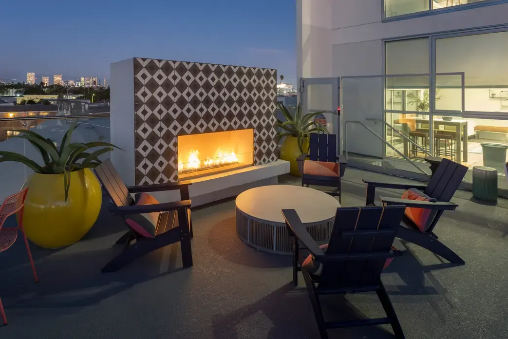 A cozy outdoor lounge area featuring a modern fire pit, surrounded by black wooden chairs and large yellow planters. The backdrop consists of a decorative patterned wall with a glowing fireplace. The setting is illuminated in twilight, showing a city skyline in the distance.