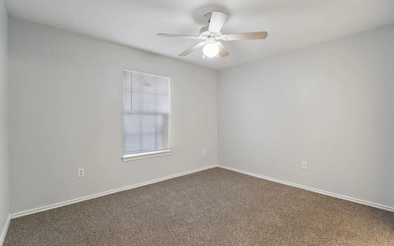 A spacious empty room with carpeted floor and a ceiling fan. There is a window with blinds on the left side of the room.