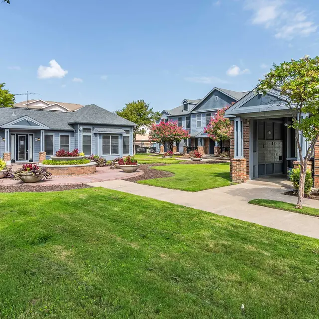 A well-maintained courtyard of an apartment complex featuring green lawns, colorful flower beds, and walkways. Buildings with gray siding and red brick exteriors are visible in the background, surrounded by trees and shrubs under a blue sky.