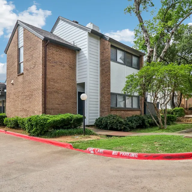 Exterior view of a two-story brick apartment building with a landscaped area and trees nearby.