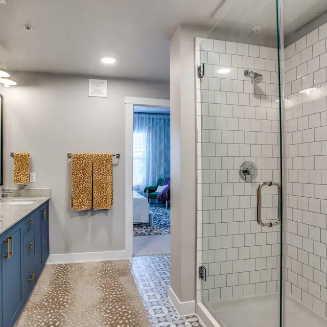 A modern bathroom featuring a glass shower, double vanity with a blue cabinet, and decorative towels. The shower has white tiled walls and patterned flooring. A glimpse of another room can be seen through an open doorway.