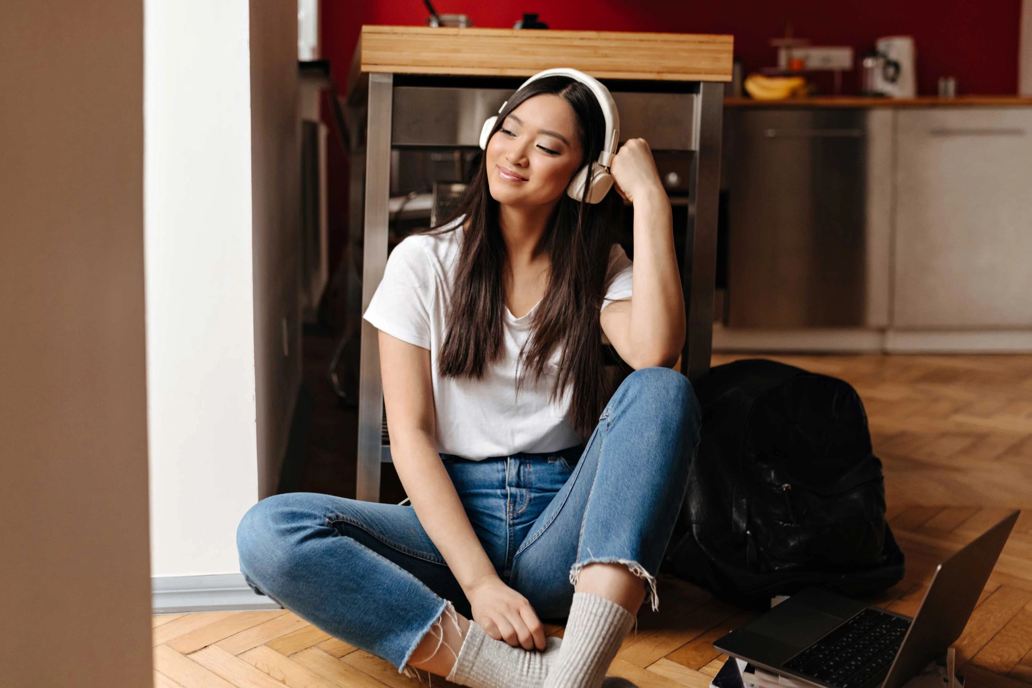 A young woman wearing headphones sits on the floor with her legs crossed, smiling gently. She is dressed casually in a white t-shirt and blue jeans. A laptop is placed next to her on the floor, and there's a black bag nearby. The background features a modern kitchen style with bright colors.