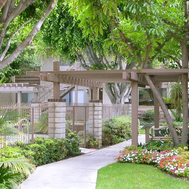 A landscaped garden pathway lined with greenery and colorful flowers, featuring a wooden pergola structure.