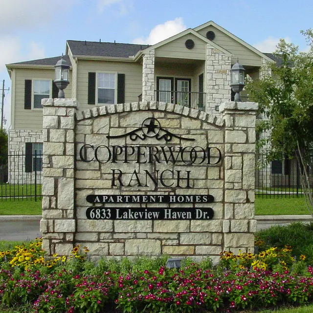 Sign for Copperwood Ranch Apartment Homes with stone facade and flowerbed in front.
