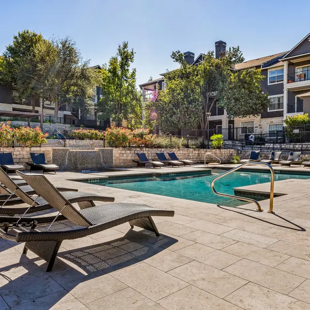 A sunny apartment complex pool area with lounge chairs and landscaped greenery.