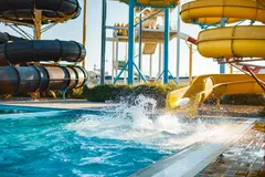 A water park with colorful water slides and a splash zone. A yellow water slide is visible, and a person is splashing into the pool from the slide. In the background, there are black tube slides and a tower with people on top.