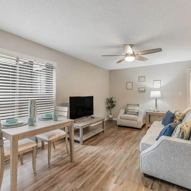 A modern and well-lit living room featuring a dining table with four chairs, a comfortable gray sofa, and an armchair. A television is positioned across from the seating area, and there are blinds on the windows allowing for natural light. The floor is wood laminate, and the walls are painted in a neutral tone.