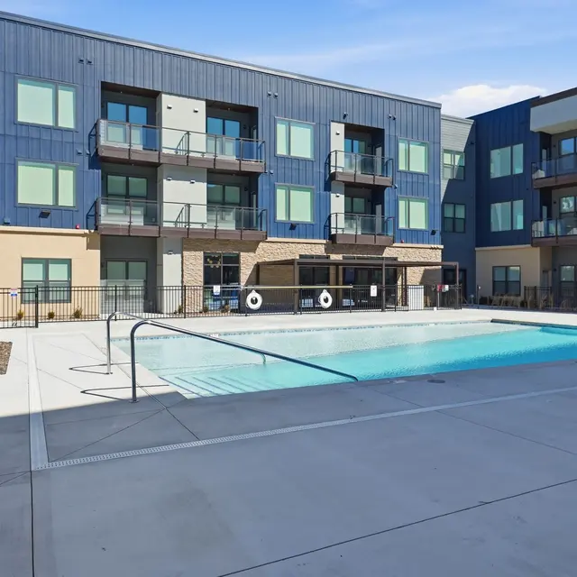 Modern apartment complex with a pool surrounded by balconies and a landscaped area. Clear blue sky enhances the scene.