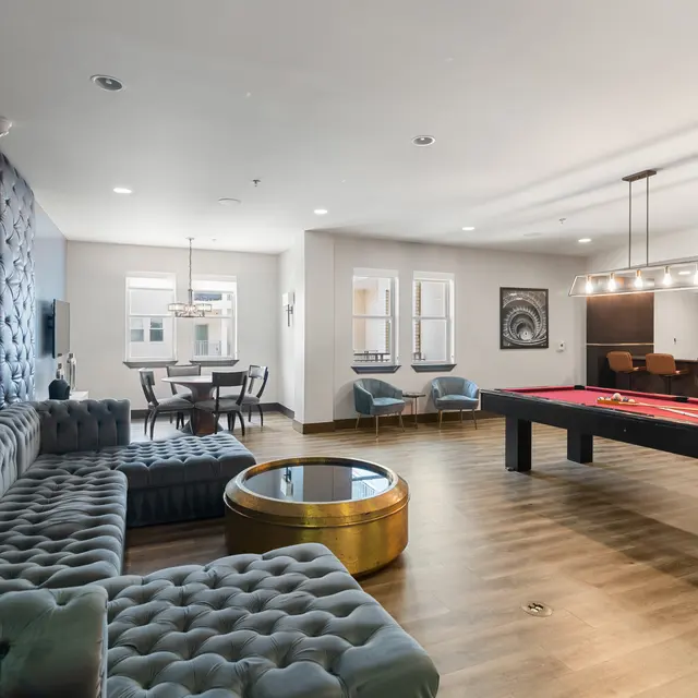 A spacious lounge area featuring a sectional sofa, pool table, dining table, and large windows allowing natural light.