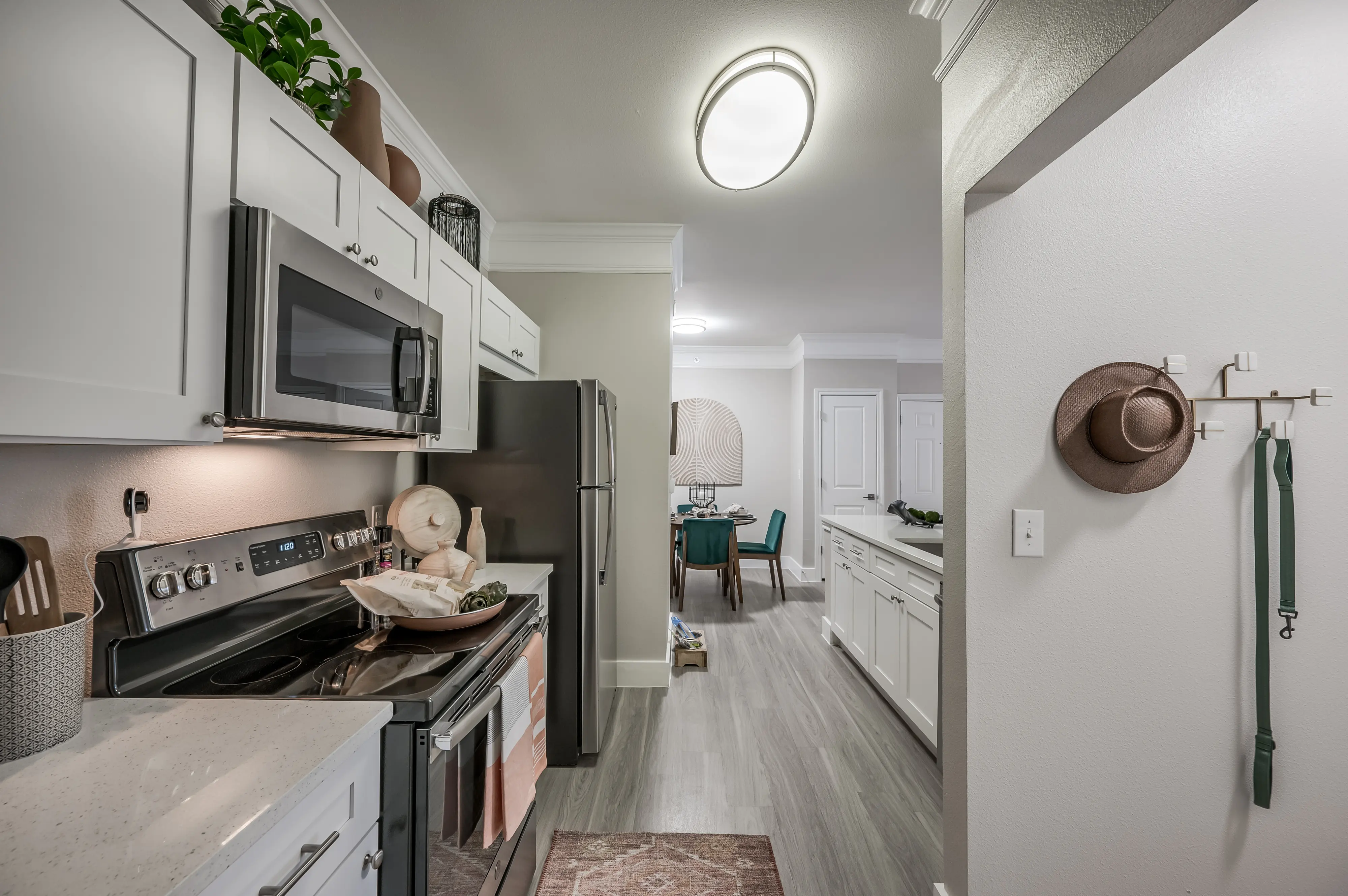 A modern kitchen featuring white cabinets, stainless steel appliances, and a neutral color palette. There's a view of a dining area in the background, with a large round table and chairs. A decorative hat is hung on the wall, adding a personal touch.