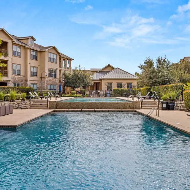 A luxurious apartment complex pool area featuring clear blue water, lounge chairs, and well-maintained landscaping.