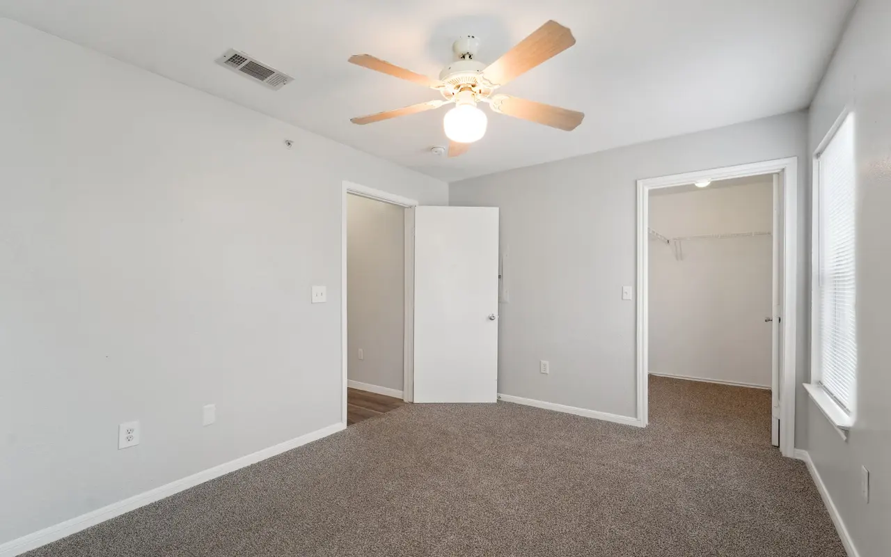 Interior view of a vacant room with gray walls and carpet, featuring a ceiling fan and two doors leading to other areas.