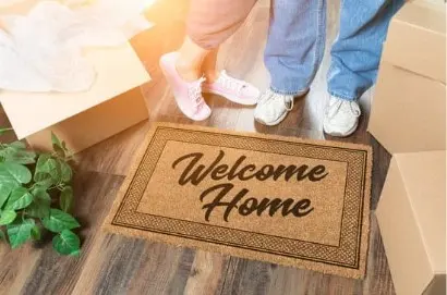 A close-up of a welcome mat that says 'Welcome Home' with two pairs of feet standing nearby, surrounded by moving boxes and a small plant on the floor.