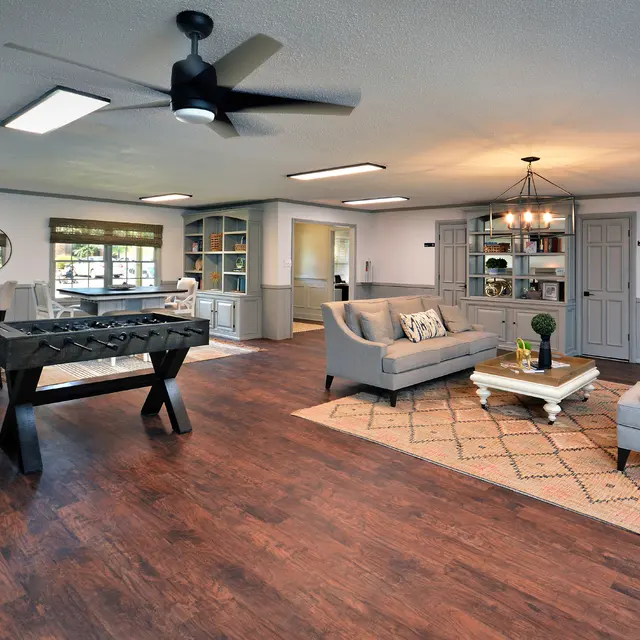 A spacious modern living room featuring a neutral color palette, a comfortable seating area with a coffee table and decorative items, a foosball table, and a dining area. The room has natural light coming through windows, wooden flooring, and stylish ceiling lights.
