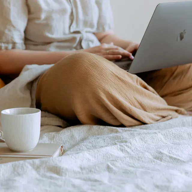 A person sitting comfortably on a bed with a laptop and a cup of coffee nearby. The person is wearing light-colored, loose-fitting clothing. A soft pillow and a blanket are visible on the bed.