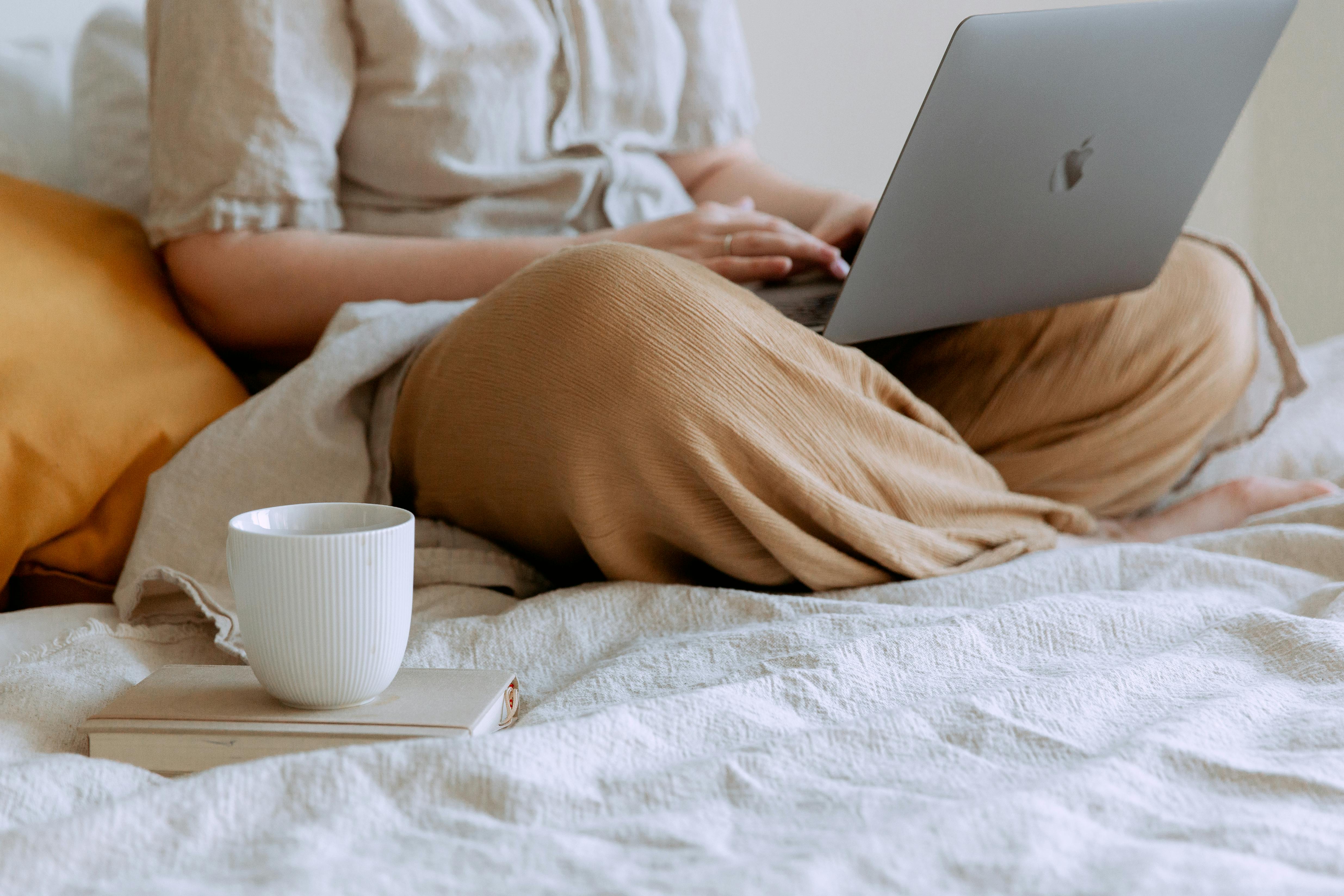Cozy Workspace at Home A person sitting comfortably on a bed with a laptop and a cup of coffee nearby. The person is wearing light-colored, loose-fitting clothing. A soft pillow and a blanket are visible on the bed.