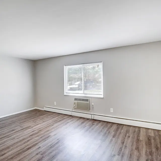 A spacious empty living room with light gray walls and brown wooden flooring. A window allows natural light to enter, and there is a wall-mounted air conditioning unit beneath the window. The room has a simple, uncluttered appearance with one door visible.