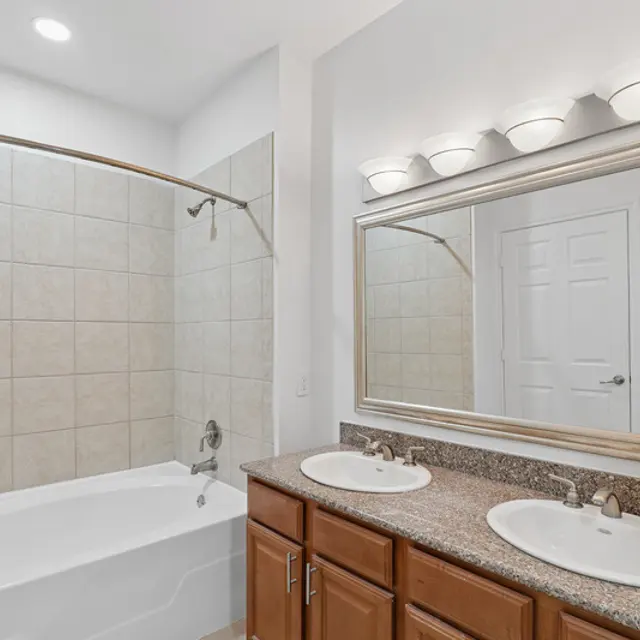 A modern bathroom featuring a curved shower rod, a bathtub, dual sinks with a granite countertop, and a large mirror with three lights above.