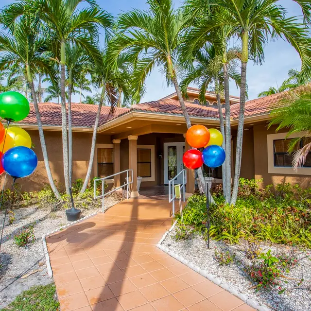 A welcoming entrance of a building surrounded by palm trees and vibrant balloons in red, orange, green, and blue.