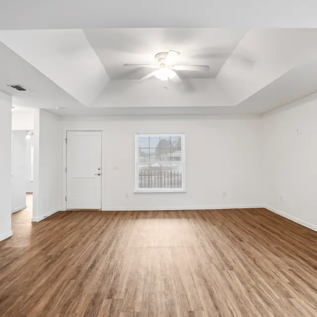 Spacious Living Room A spacious and empty living room featuring wooden flooring, white walls, a ceiling fan, and a window. The room has a doorway leading to another area and an open layout.