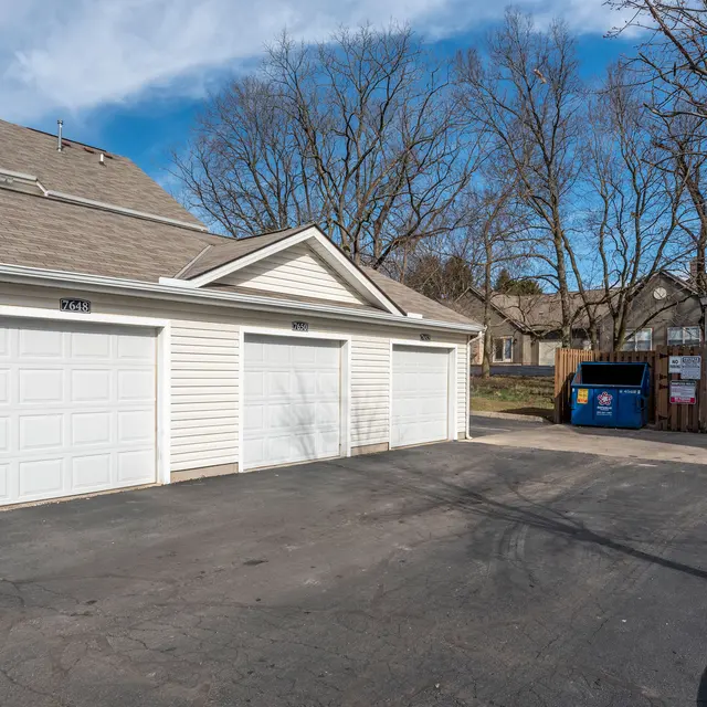A residential garage with three doors and a driveway, adjacent to two blue dumpsters. The area is sunny with trees in the background and a few houses visible.