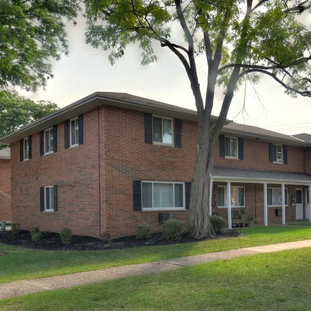 A brick residential apartment building with multiple units, surrounded by grassy areas and trees.