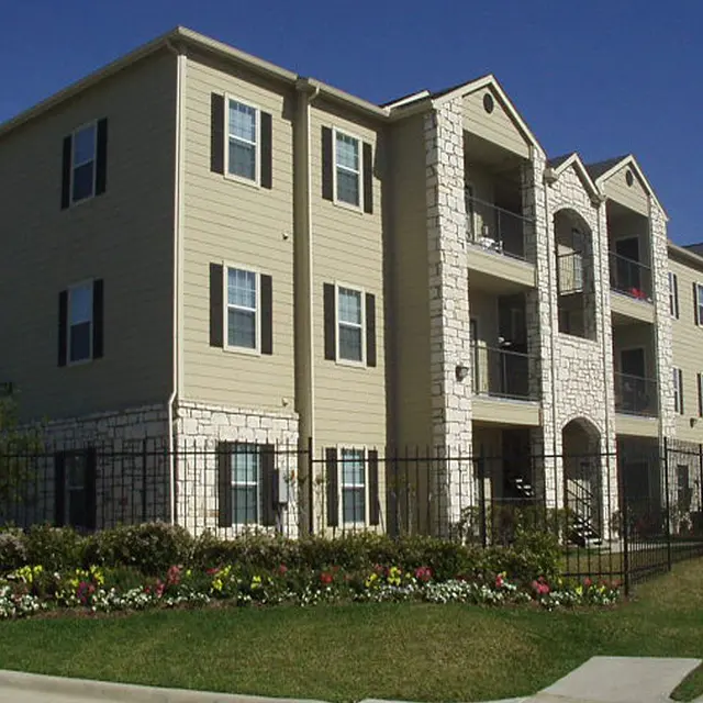 A modern apartment building featuring stone and beige siding, with balconies and large windows, surrounded by a landscaped garden.