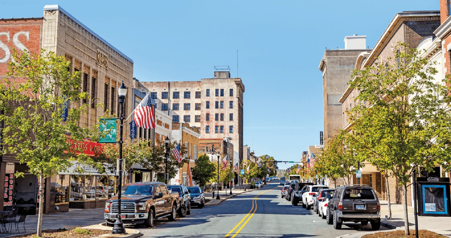 A vibrant downtown street lined with trees, featuring a mix of historic buildings and modern storefronts. Cars are parked along the street, and American flags are displayed on several buildings under a clear blue sky.