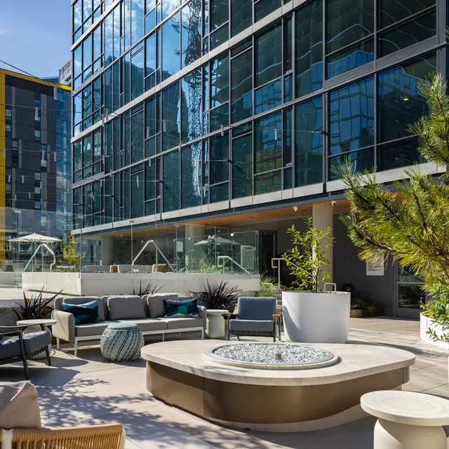 A stylish outdoor lounge area featuring comfortable seating, potted plants, and a circular fire pit, alongside a modern glass building.