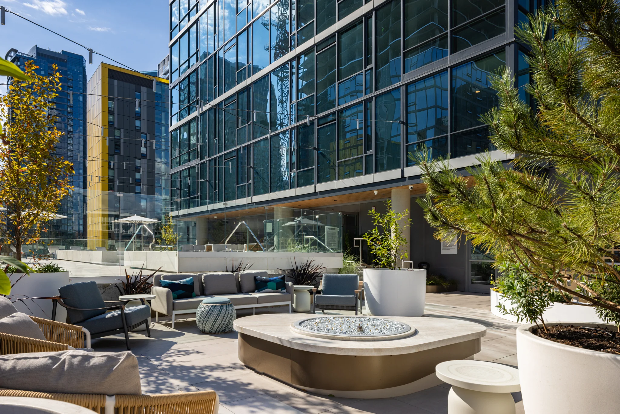 A stylish outdoor lounge area featuring comfortable seating, potted plants, and a circular fire pit, alongside a modern glass building.