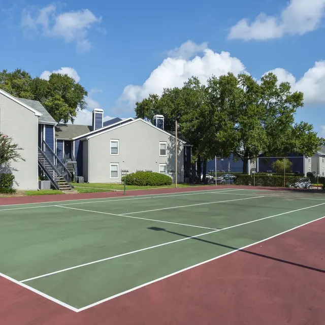A sunny tennis court surrounded by apartment buildings and lush trees under a blue sky with fluffy clouds.