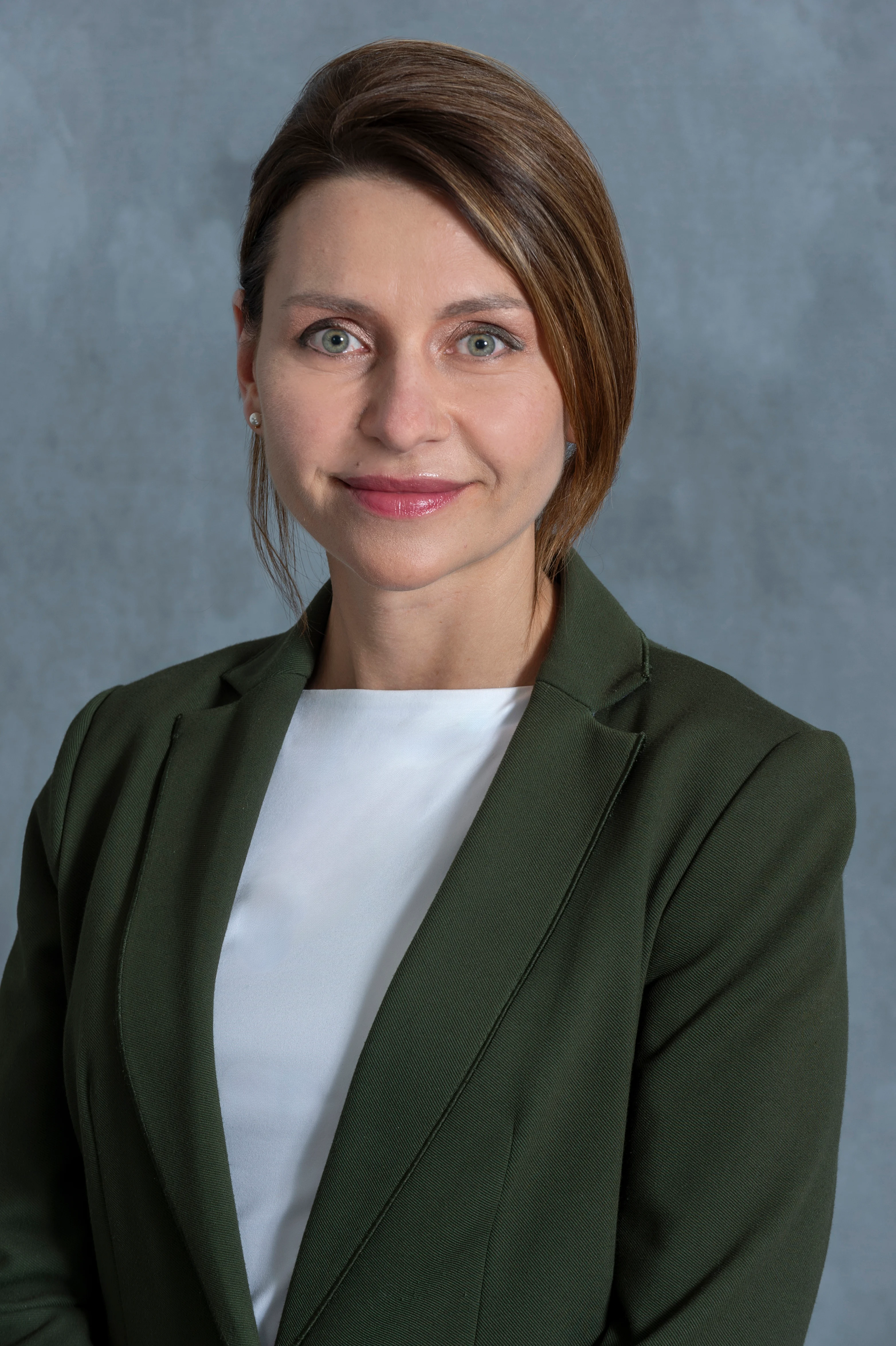 A woman in a professional green blazer with a white blouse, smiling softly against a light gray background.