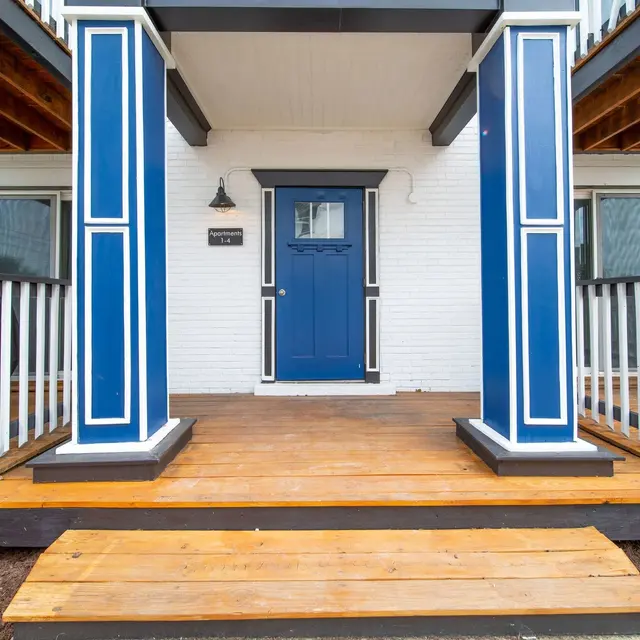 A front porch with wooden steps leading to a blue door, flanked by white columns and railings.
