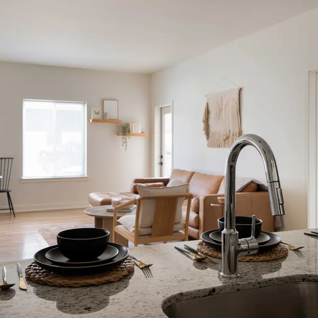 A modern, open-plan living space featuring a light-filled area with a cozy sofa, dining table, and chairs. The kitchen is visible in the foreground with a granite countertop and dishware set up for a meal.