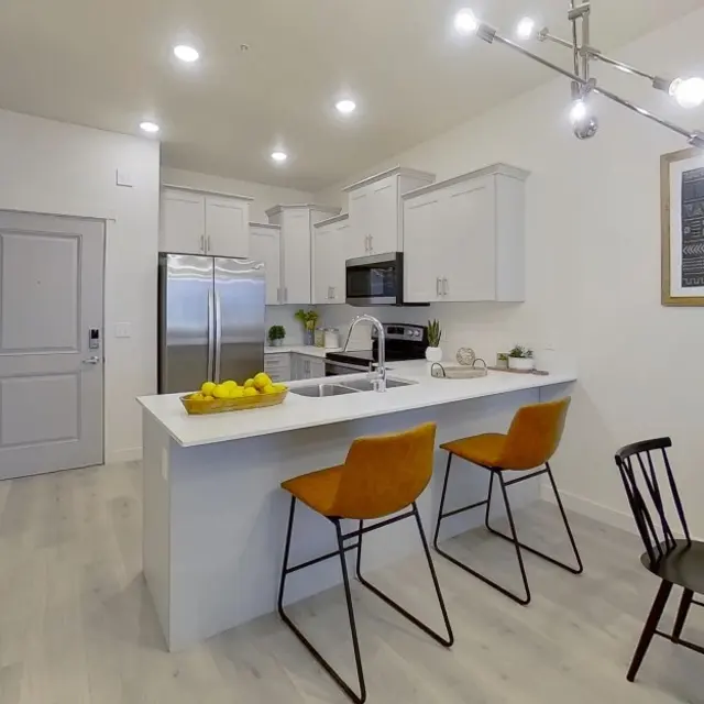 Modern Kitchen Design A modern kitchen featuring white cabinets, stainless steel appliances, and a light countertop. There are two orange bar stools at a small kitchen island and a dining area with a round table and chairs.