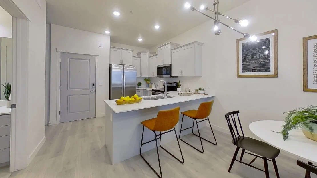 A modern kitchen featuring white cabinets, stainless steel appliances, and a light countertop. There are two orange bar stools at a small kitchen island and a dining area with a round table and chairs.
