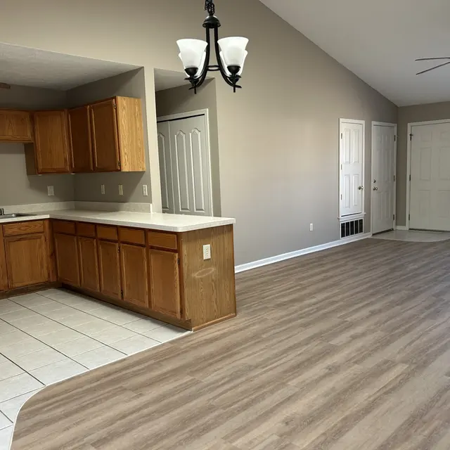An open-concept living space featuring a kitchen with wooden cabinets and a dishwasher on the left, and a spacious living area with wooden flooring and a ceiling fan.